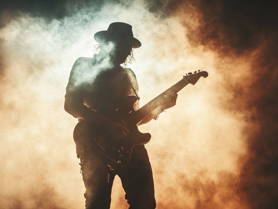 Silhouette of a guitarist wearing a hat, performing under smoky stage lighting—capturing the essence of live rock music energy.