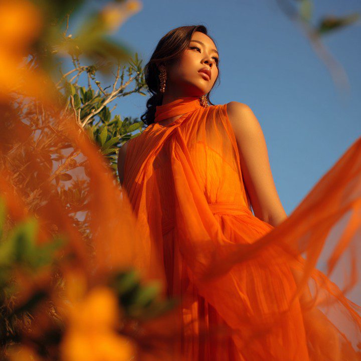 Stylish woman wearing a flowing orange georgette dress in sunlight, posing outdoors with natural greenery and sky background.