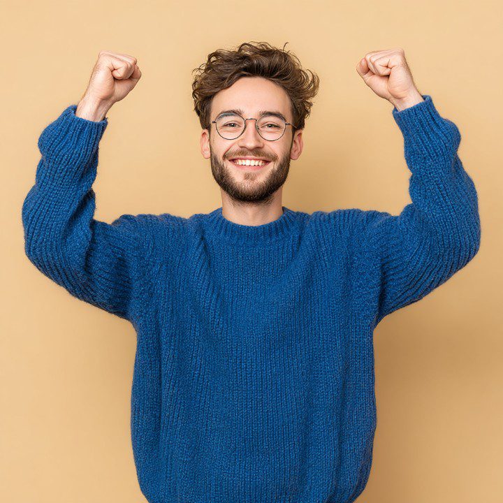 A happy young man wearing glasses and a blue sweater raises his arms in triumph against a beige background.