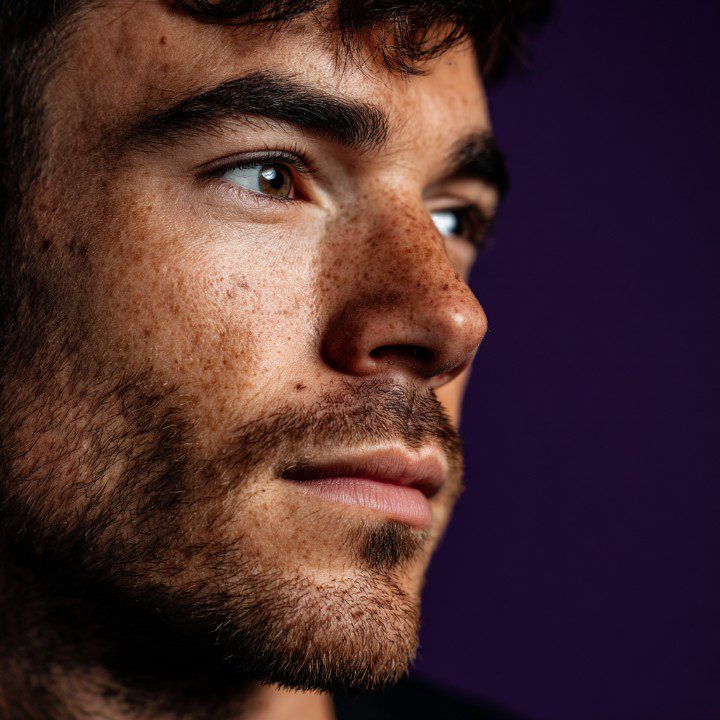 Close-up of a bearded man looking away thoughtfully with a purple background, emphasizing emotion and texture.