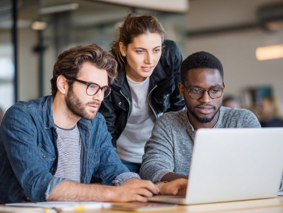Three diverse professionals collaborating on a laptop in a modern office, representing teamwork and innovation.