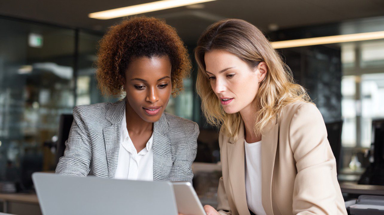 Two diverse businesswomen collaborate using a laptop and tablet in a modern office, symbolizing teamwork and digital innovation.
