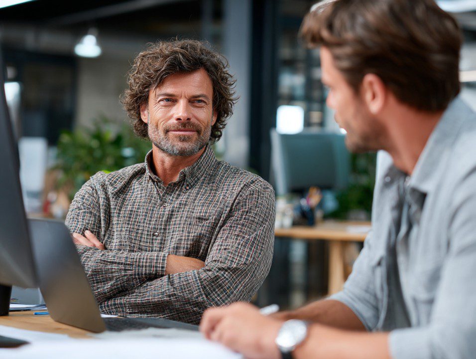 Middle-aged office manager listening to a colleague at a desk in a modern office, suggesting leadership or mentoring.
