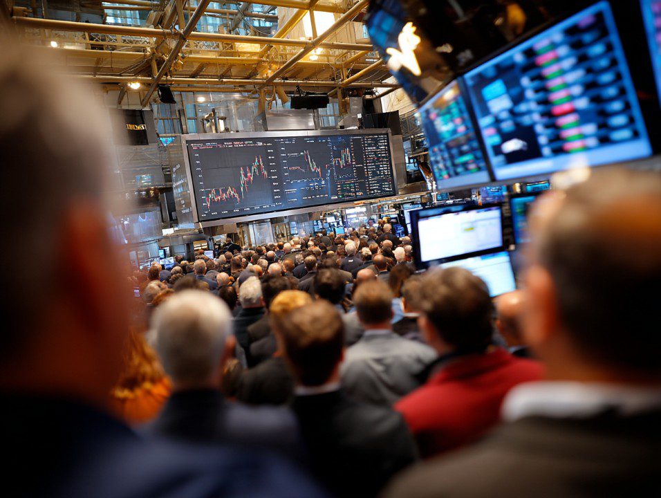 Crowded trading floor with people watching large screens displaying live financial charts and stock market data.