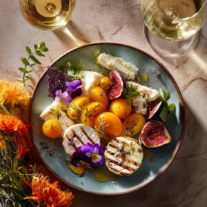 Top-down view of a gourmet plate with grilled cheese, figs, yellow tomatoes, and edible flowers, paired with white wine.