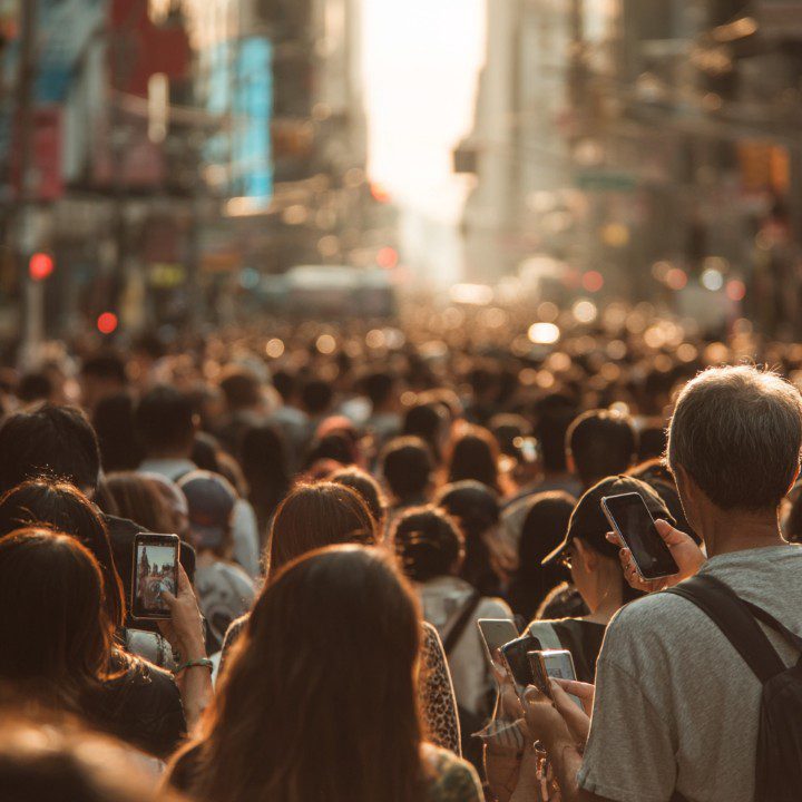 A dense crowd of people in a city street at sunset, most of them using smartphones, reflecting modern digital behavior.