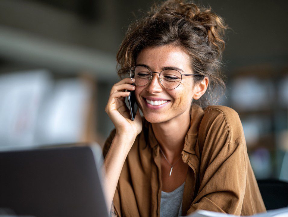 Happy businesswoman wearing glasses talking on the phone and smiling while working on a laptop in a casual office.