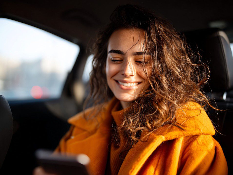 Cheerful young woman in orange coat smiles at her smartphone while seated in a car, representing digital connectivity and lifestyle.