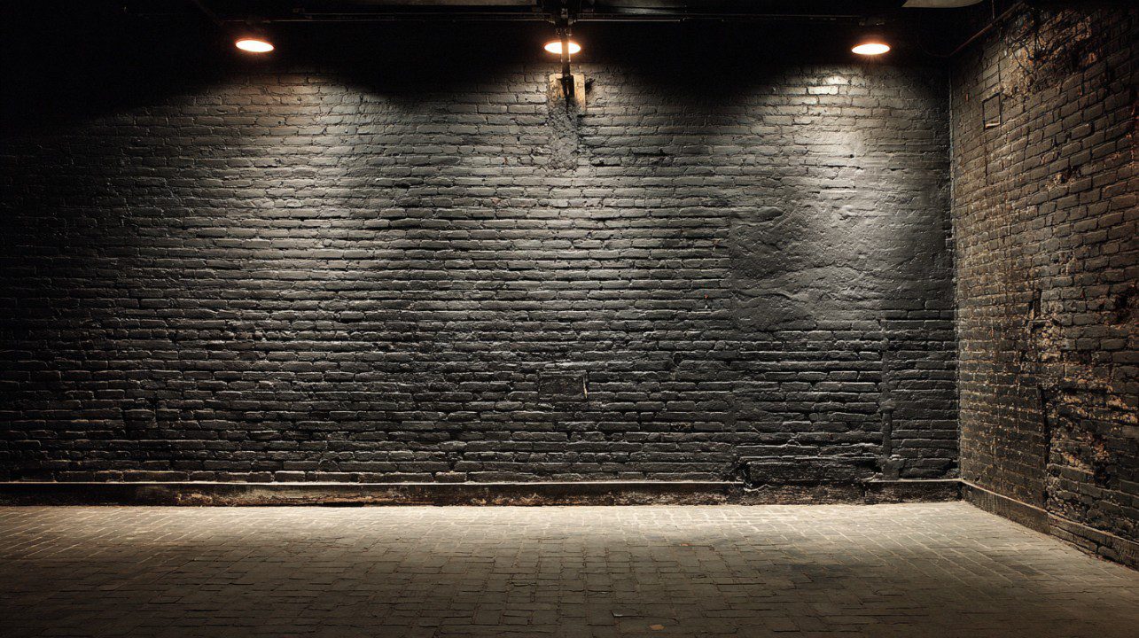 Black painted brick wall illuminated by three overhead lights in a dark theater or room, creating a moody industrial atmosphere.