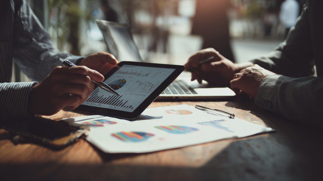 Two professionals analyzing financial charts on a tablet and paper in a sunlit meeting, symbolizing strategy and collaboration.