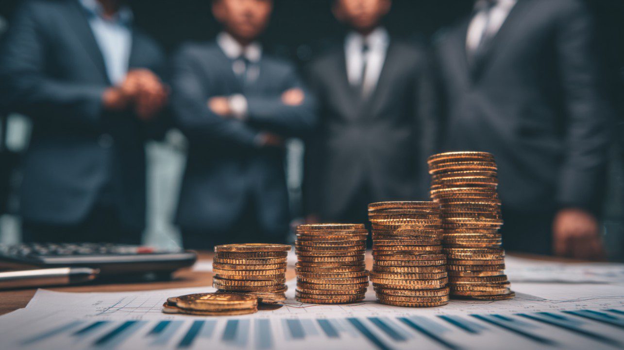 Business team in suits analyzing stock market investment with stacks of gold coins on financial charts.