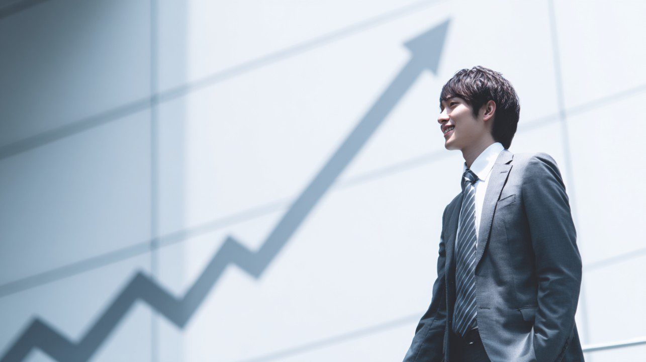 A confident businessman in a suit smiles while standing beside a wall with a rising arrow, symbolizing business growth and success.