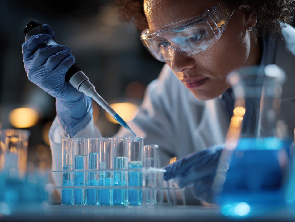 Female scientist wearing goggles and lab coat carefully using a pipette to transfer blue liquid in test tubes.