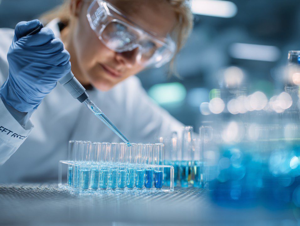 Female scientist in lab coat and safety goggles transferring blue liquid into test tubes using a pipette.