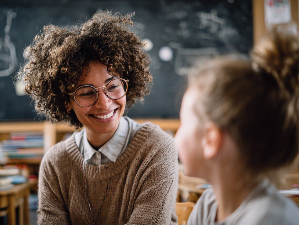 A cheerful teacher with glasses warmly smiles at a young student in a cozy classroom, illustrating supportive learning.