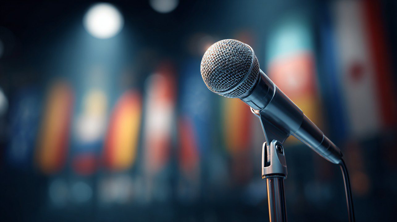 A close-up of a microphone lit by a spotlight with blurred international flags in the background, ready for a global address.