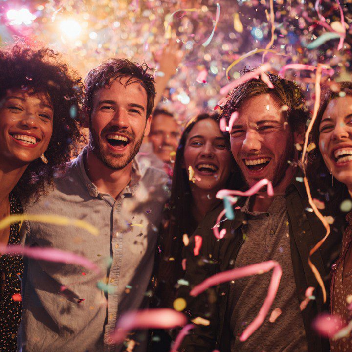 A group of friends smiling and laughing during a night party, surrounded by falling confetti and vibrant lighting.