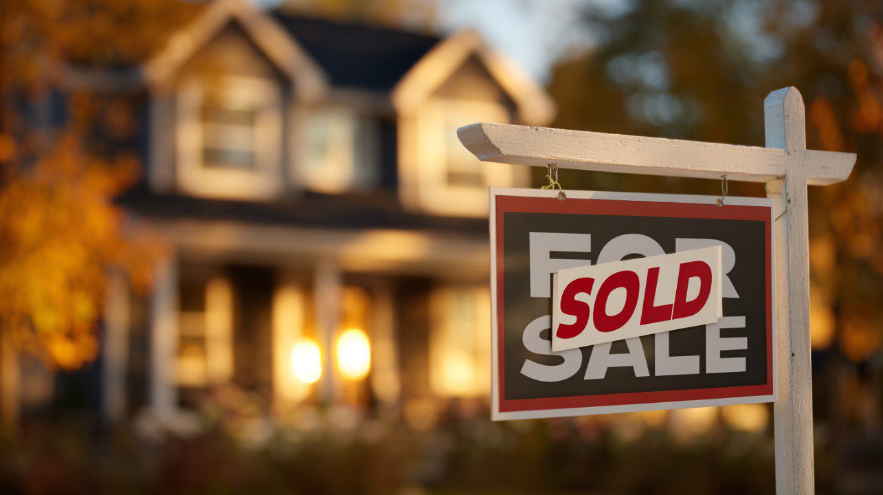 Close-up of a real estate "For Sale" sign with a red "Sold" sticker, blurred suburban house in background.
