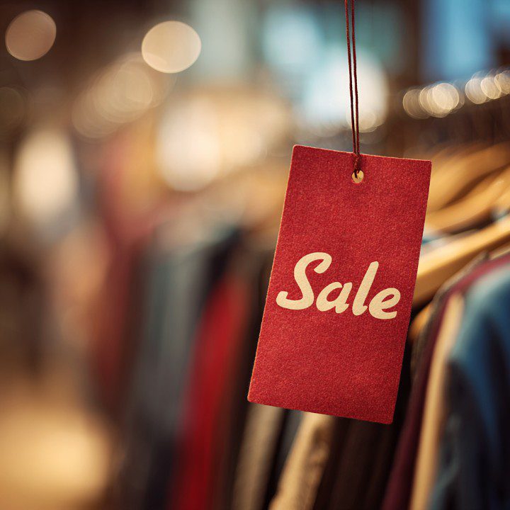 Close-up of a red sale tag hanging on a clothing rack with blurred clothing and store lights in the background.