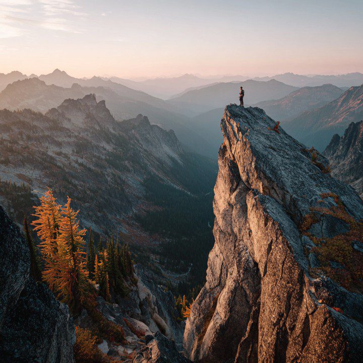 A lone rock climber stands on a mountain peak at sunrise, overlooking distant mountain ranges.