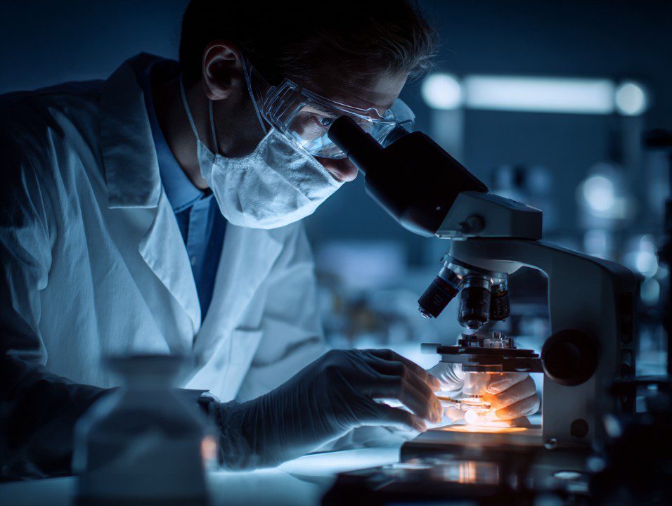 Scientist wearing mask and gloves observing a sample under a microscope in a modern laboratory setting.