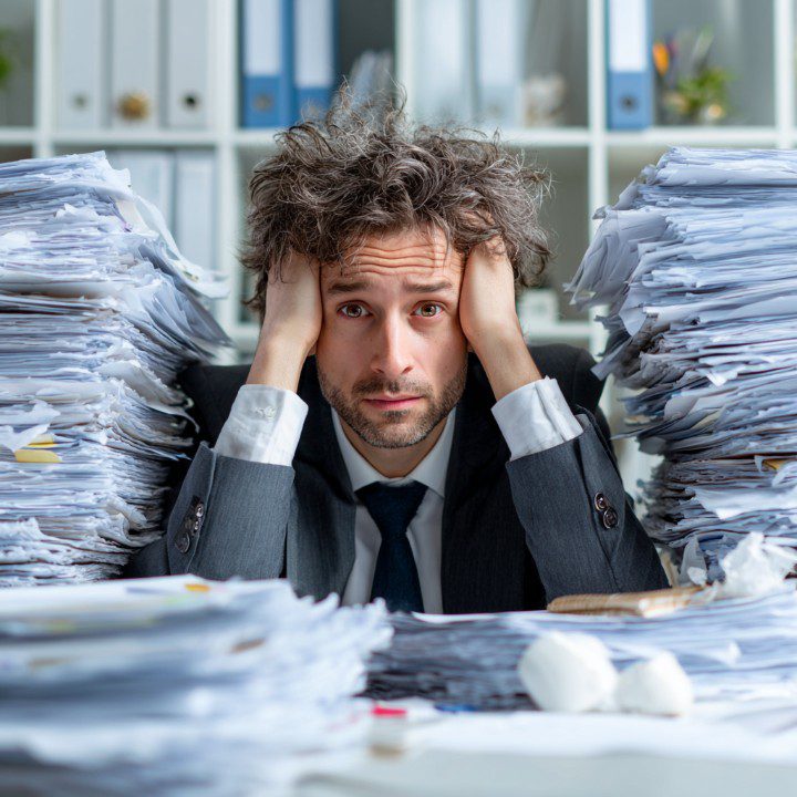 A stressed business manager in a suit overwhelmed by towering piles of paperwork at his office desk.