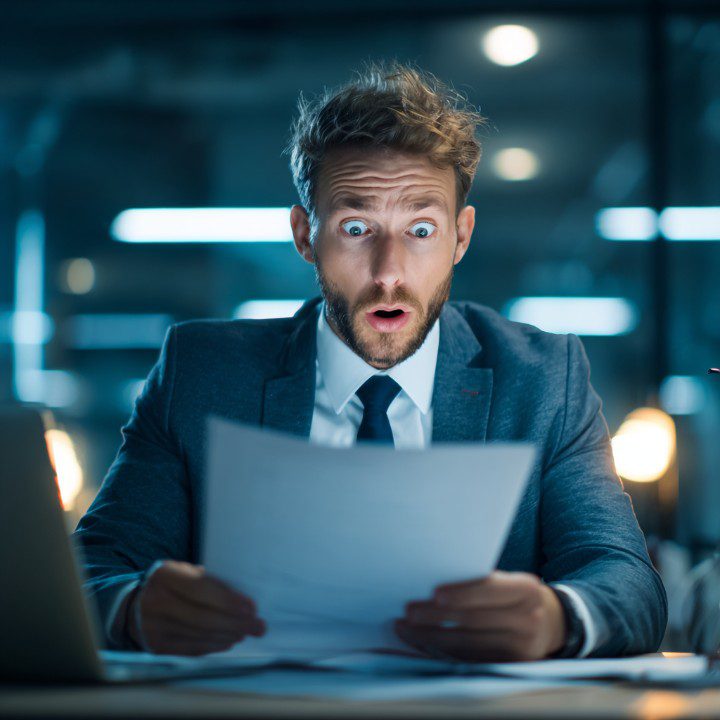 Businessman in office suit showing shocked expression while reading documents at his desk.