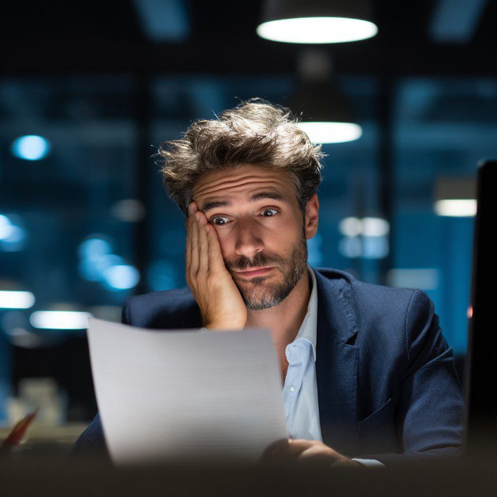 Businessman in office suit looking stressed while reviewing papers in dimly lit office.
