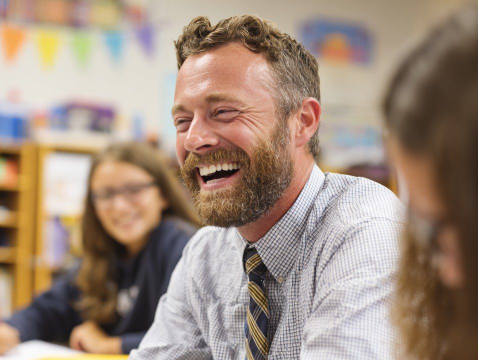 Teacher with beard laughing joyfully in classroom with students blurred in the background.