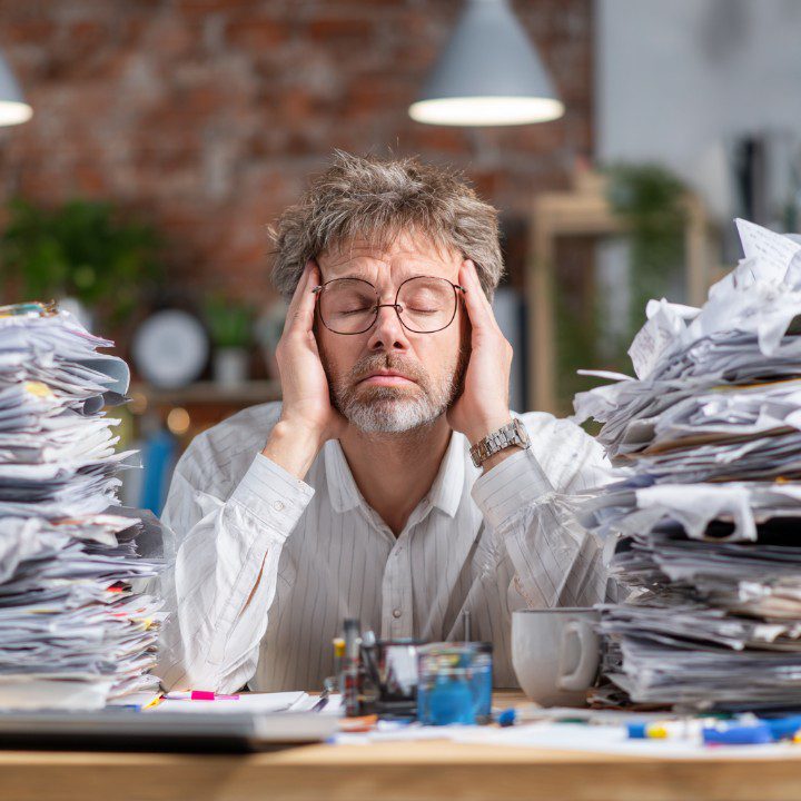 A tired business manager with glasses sits at a desk overwhelmed by tall stacks of paperwork in an office.