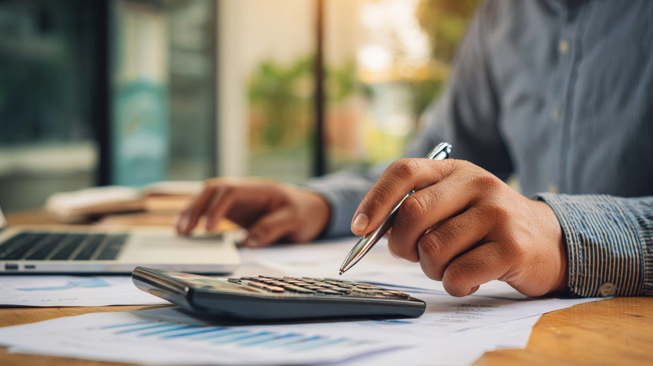 Person using a calculator and laptop while reviewing financial documents in an office.