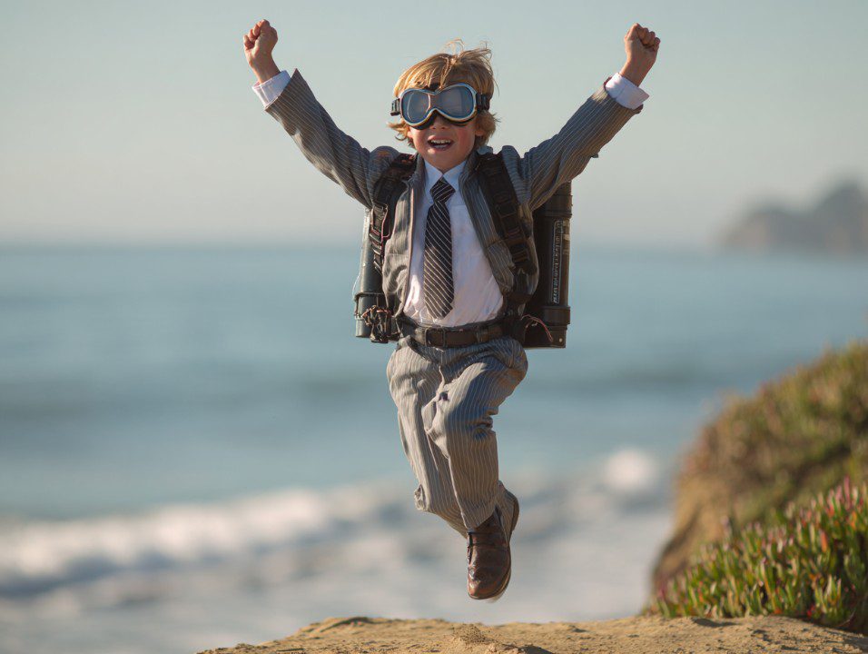 Young boy in a suit and tie with a jet pack and aviator goggles jumping with joy by the ocean.