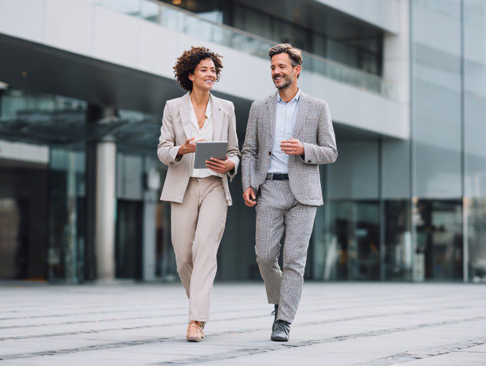Two business professionals walking and using a digital tablet in a modern city setting.