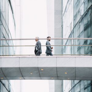 Two businessmen in suits walking across a modern elevated walkway between office buildings.