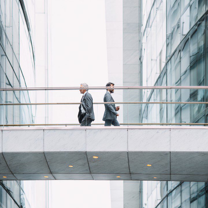 Two businessmen in suits walking across a modern elevated walkway between office buildings.