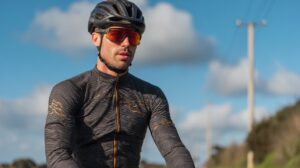 Male cyclist wearing a black patterned cycling jersey, helmet, and red-tinted sunglasses during an outdoor ride under a sunny blue sky.
