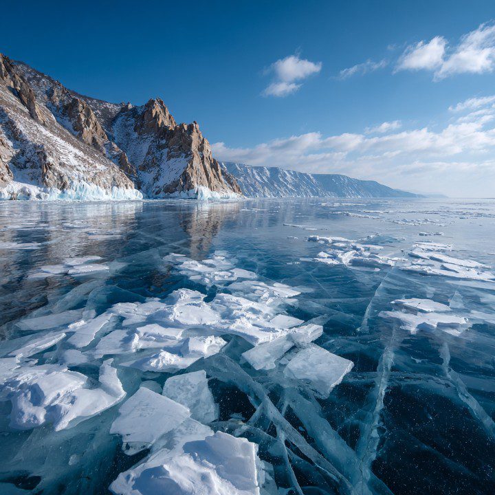 Frozen Lake Baikal with cracked ice and snow-covered cliffs under a blue sky.
