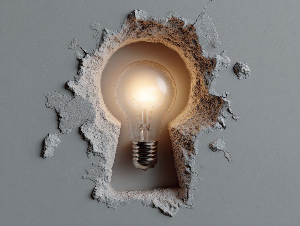 Glowing light bulb in keyhole-shaped break in a wall, representing innovation and problem-solving.