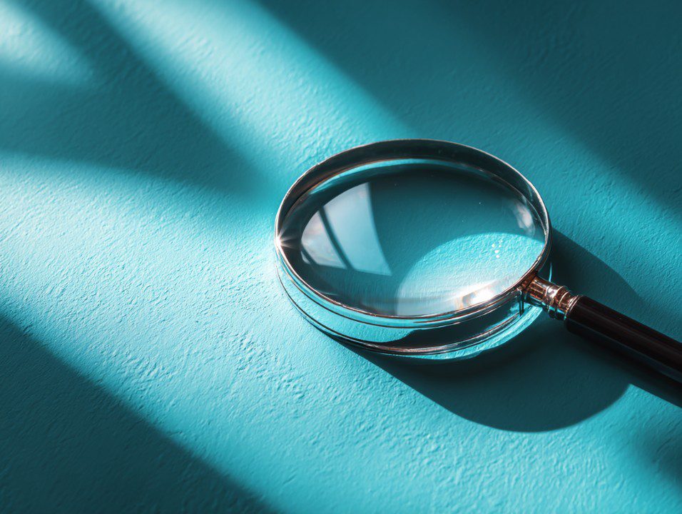 Close-up magnifying glass resting on a turquoise background with soft light and shadows.