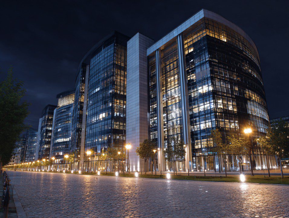 Modern glass office building in Paris illuminated at night with cobblestone street and streetlights.