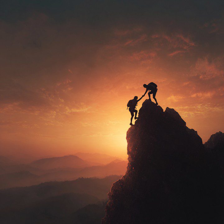 Silhouette of two climbers at sunrise, one helping the other to the mountain peak.