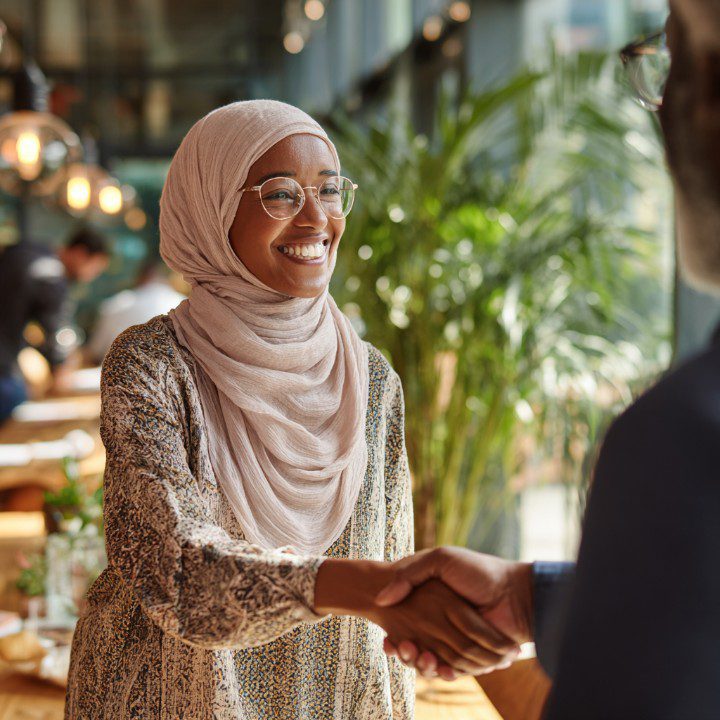 Professional Muslim woman wearing a light hijab smiling while shaking hands in a bright office.