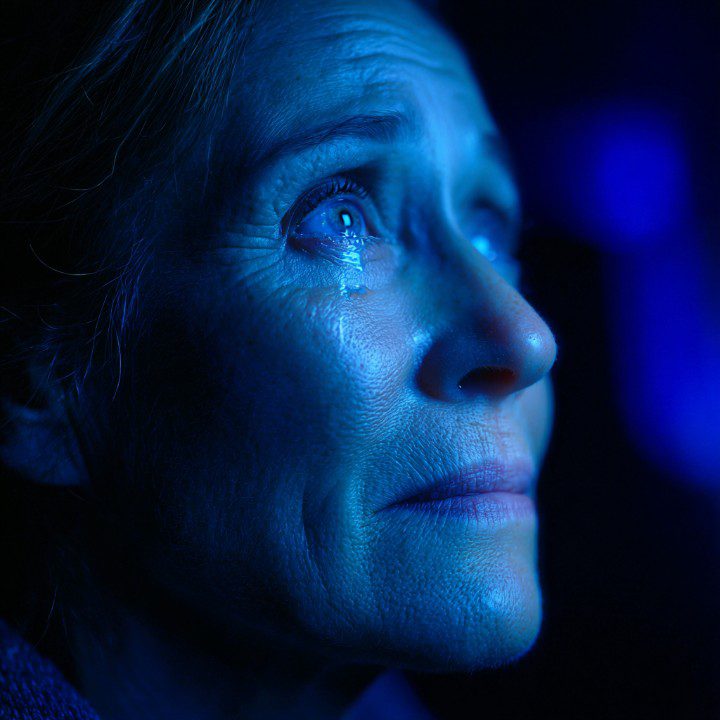 Older woman with tears in her eyes illuminated by blue light, looking up with emotion.