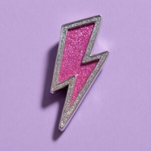Close-up of a pink glitter lightning bolt pin outlined in silver on a lavender background.