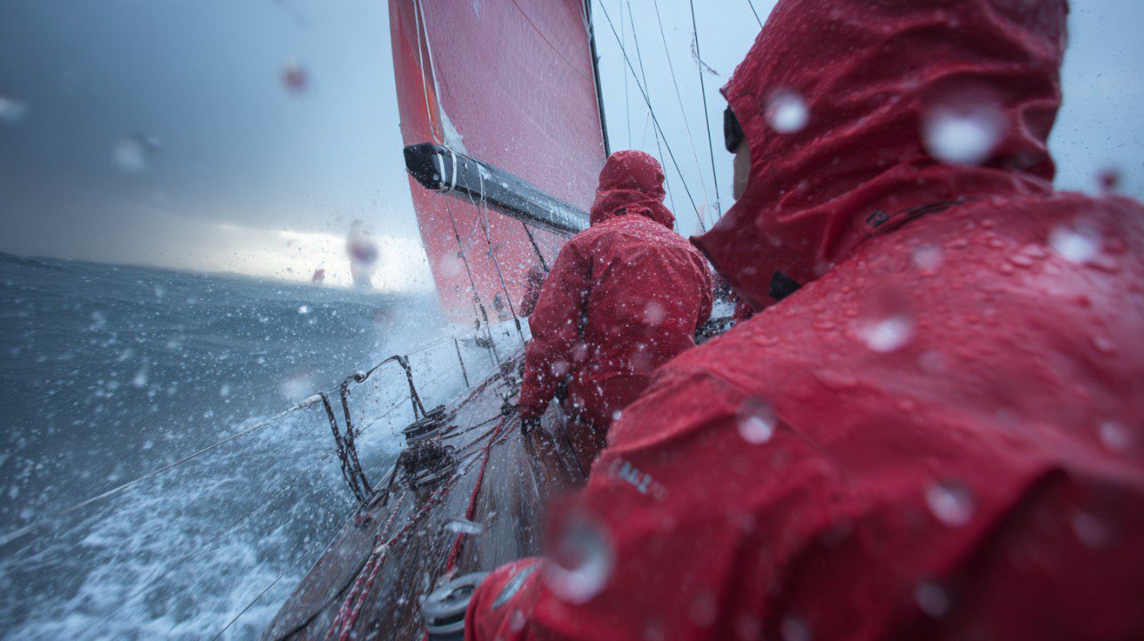 Racing yacht crew in red waterproof jackets sailing through rough seas with water splashing over the deck.