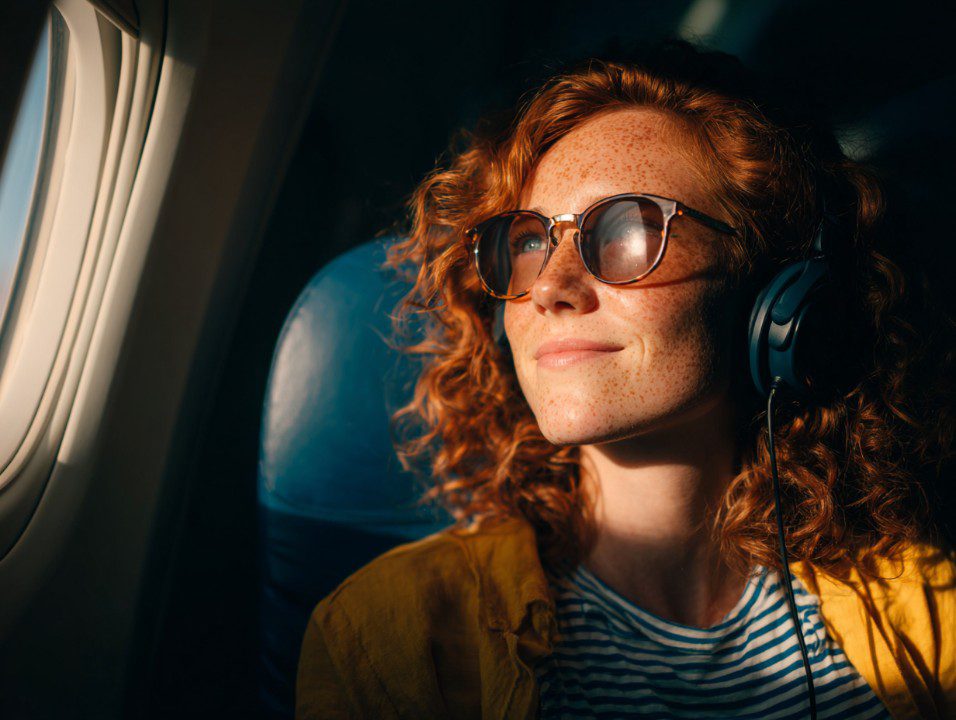 Red-haired woman with freckles, sunglasses, and headphones looking out of an airplane window during golden hour.