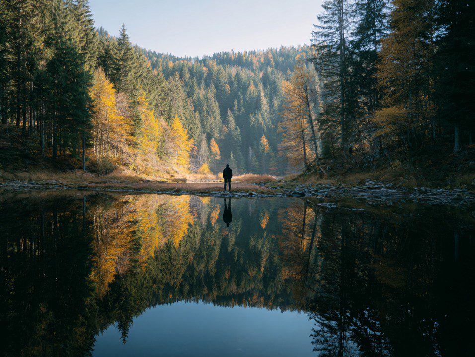 A lone person stands at the edge of a calm lake with autumn trees reflected in the water.