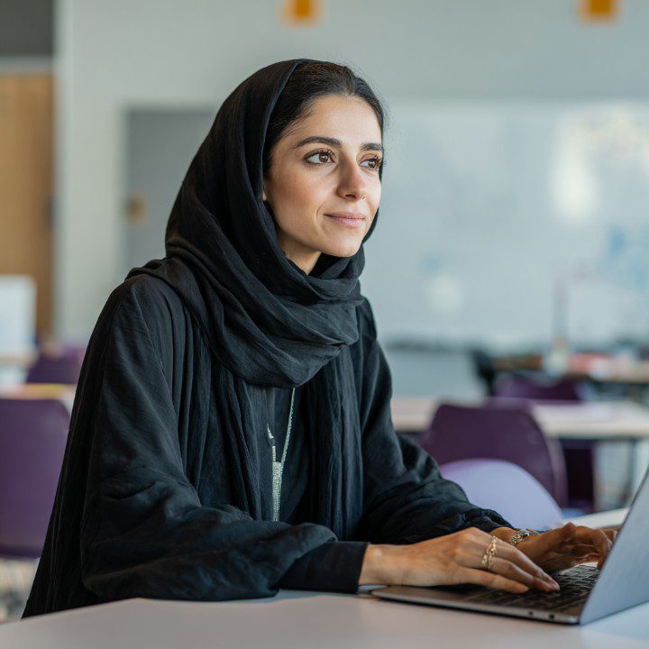 A Saudi female teacher in a black abaya and hijab works on a laptop in a modern classroom setting, symbolizing education and professionalism.