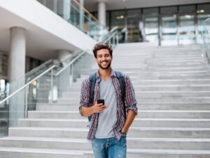 Cheerful male student with a backpack and smartphone standing on wide modern university stairs in casual jeans and a plaid shirt.