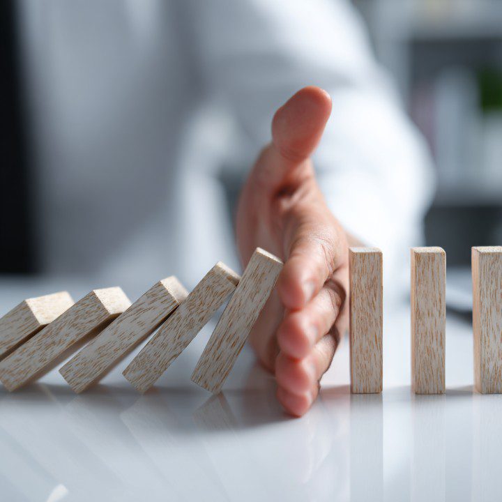 A close-up of a hand stopping wooden domino blocks from falling, symbolizing risk management, prevention, and crisis control.